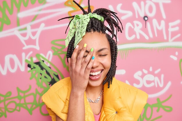 Веселая многолетняя девушка искренне смеется, делает лицо ладони очень счастливой, имеет модную прическу, одетую в повседневную одежду, позирует на фоне красочной стены с граффити.