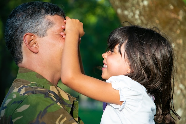 Free photo cheerful military father holding little daughter in arms while joyful girl shutting his and smiling. side view. family reunion or returning home concept