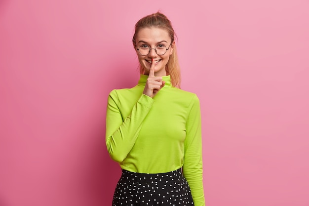 Cheerful millennial girl prepares surprise for someone smiles tenderly makes silence gesture asks keep secret wears optical glasses casual turtleneck 