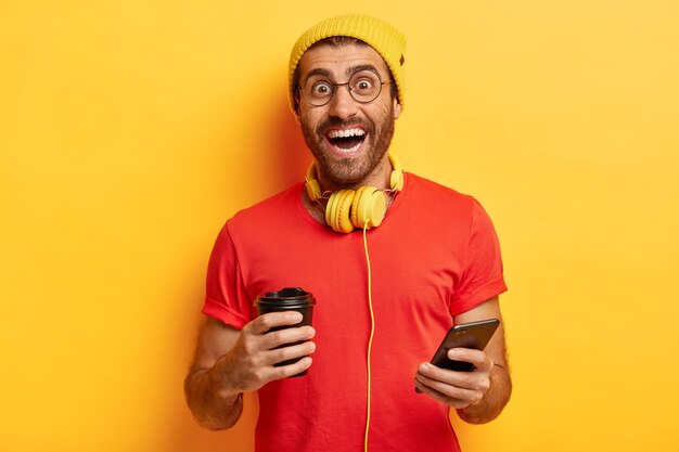 Cheerful millennial guy stands with coffee cup and smartphone