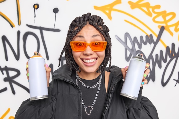 Cheerful millennial woman with braids holds two aerosol spray bottles being creative street artist draws graffiti wears fashionable clothes orange sunglasses belongs to gang