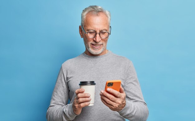 陽気な現代の年配の男性は、通信の種類に携帯電話を使用しています電話画面のテキストメッセージは青い壁に隔離されたカジュアルな服を着たコーヒースクロールインターネットページの紙コップを保持します