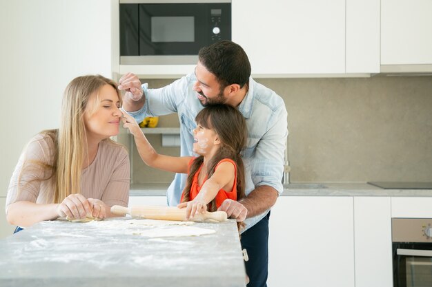 Cheerful mom, dad and girl staining faces with flower powder while baking together.