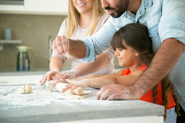 Cheerful mom and dad teaching happy daughter to roll dough on kitchen table with flour messy. young couple and their girl baking buns or pies together. family cooking concept