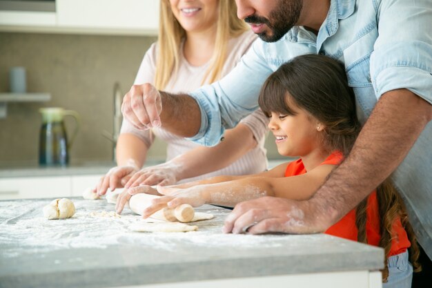Cheerful mom and dad teaching happy daughter to roll dough on kitchen table with flour messy. Young couple and their girl baking buns or pies together. Family cooking concept