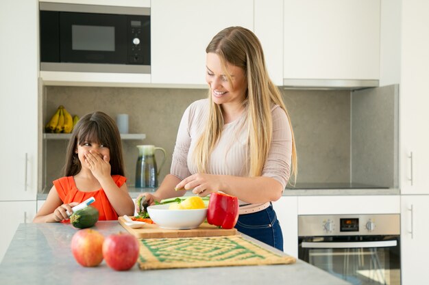 陽気なママと娘が夕食に野菜を作りながらおしゃべりして笑っています。少女と彼女の母親は、キッチンカウンターでサラダの野菜を剥離し、切断します。家族の料理のコンセプト