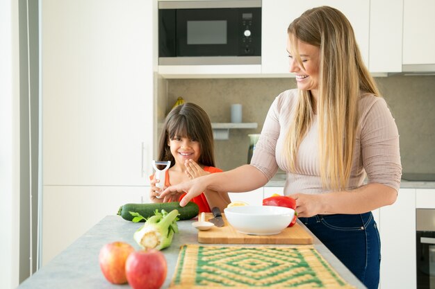 Cheerful mom and daughter having fun while cooking vegetables for dinner. Girl and her mother peeling and cutting vegs for salad on kitchen counter, chatting and laughing. Family cooking concept