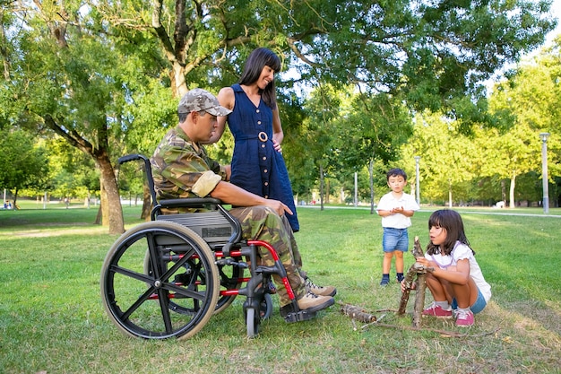 Cheerful mom and disabled military dad in wheelchair looking at kids arranging firewood for campfire outdoors. Disabled veteran or family outdoors concept