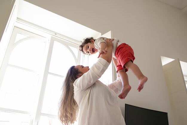 쾌활 한 엄마 팔에 흥분된 아기를 들고 공기에 손을 올려. 낮은 각도. 가정 및 어린 시절 개념에서 아이