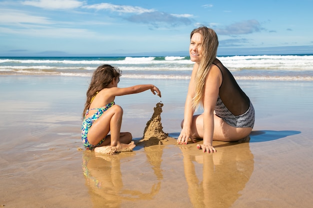 Cheerful mom and little daughter building sandcastle on beach, sitting on wet sand, enjoying vacation at sea
