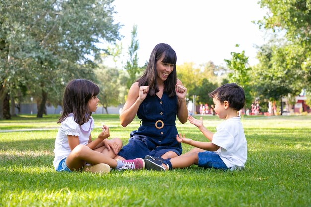 Cheerful mom and two kids sitting on grass in park and playing. Happy mother and children spending leisure time in summer. Family outdoors concept