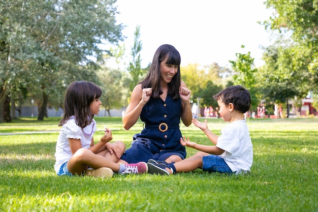 Cheerful mom and two kids sitting on grass in park and playing. Happy mother and children spending leisure time in summer. Family outdoors concept