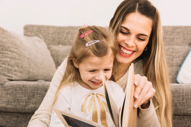 Free photo cheerful mother and daughter reading book near sofa