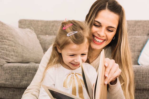 Cheerful mother and daughter reading book near sofa