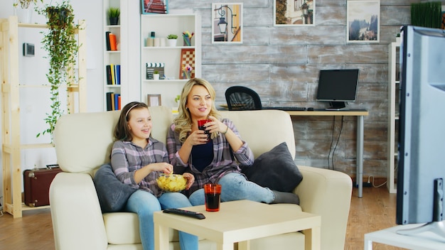 Веселая мать и дочь, сидя на диване в гостиной, смотрят телевизор, едят чипсы и питьевую газировку.