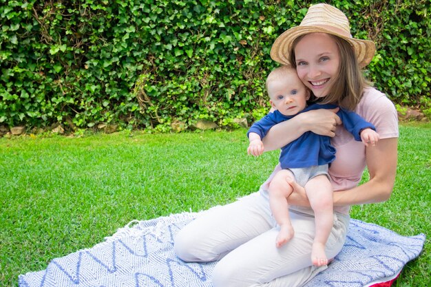 Cheerful mother in hat sitting on plaid with cute newborn in park and holding him. Little red-haired baby with bare legs looking at camera. Front view