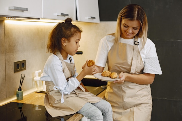 Madre allegra e figlia piccola che mangiano biscotti appena sfornati in cucina, si godono la pasticceria fatta in casa, indossano grembiuli e si sorridono a vicenda, divertendosi a casa.