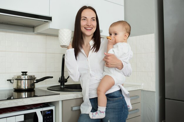 Cheerful mother standing in kitchen, drinking morning coffee and holding baby