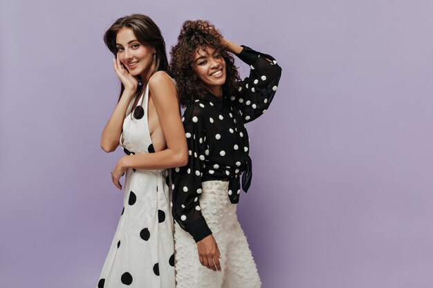 Cheerful mulatto lady in polka dot black shirt and stylish pants smiling and posing with charming girl in modern white clothes