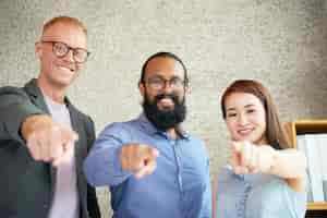 Free photo cheerful multiethnic colleagues standing in office and pointing towards camera