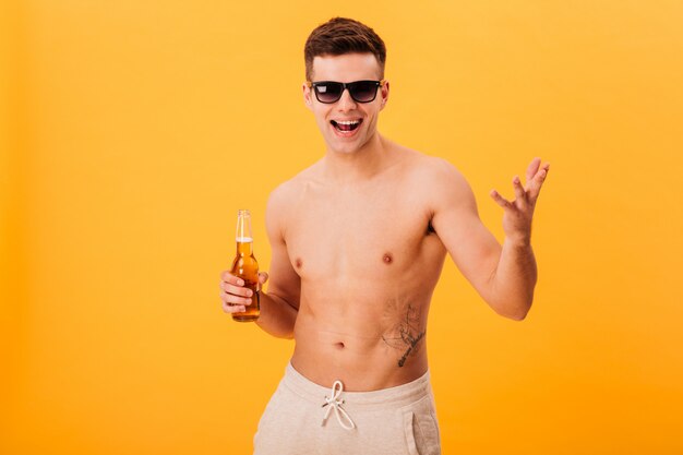 Cheerful naked man in shorts and sunglasses holding bottle of beer 