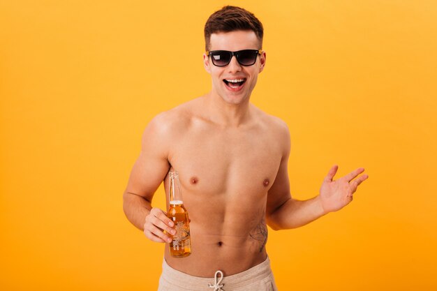 Веселый голый мужчина в шортах и солнцезащитных очках держит бутылку пива