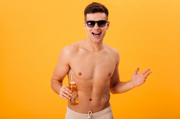 Cheerful naked man in shorts and sunglasses holding bottle of beer 