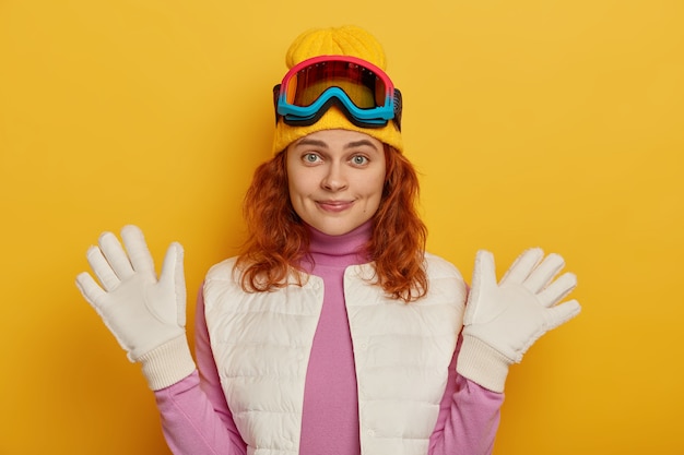 Cheerful natural woman raises hands in white gloves, wears snowboard glasses, enjoys sunny winter day, looks happily at camera, poses against yellow background.