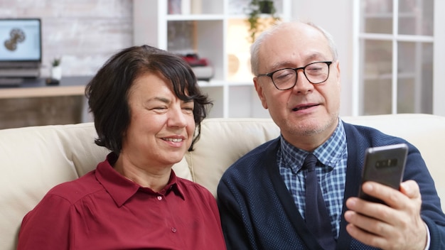 Cheerful old couple sitting on sofa taking a selfie in living room.