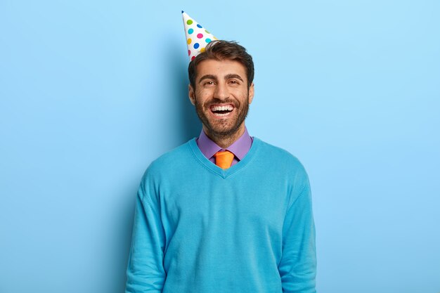 青いセーターでポーズをとる誕生日の帽子を持つ陽気な楽観的な男