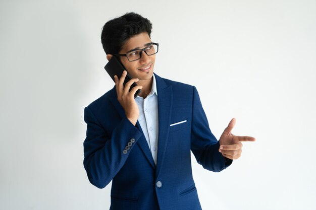 Cheerful optimistic Indian businessman talking on mobile phone and making finger gun