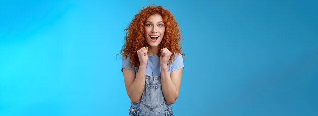 Cheerful optimistic lucky redhead curlyhaired attractive woman cheering clench fists joyfully smilin