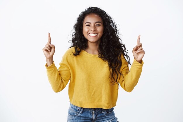 Cheerful optimsitic young female student with curly dark hairstyle, yellow sweater, smiling and laughing happily, pointing fingers up, showing friends link to site or copy space, white wall