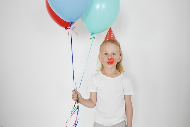 Cheerful overjoyed schoolboy with blond hair wearing holiday cone hat and white t-shirt posing at white blank wall, holding blue and red balloons, blowing whistle, having fun at birthday party