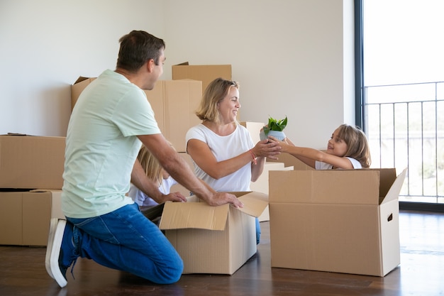 Веселые родители и дети распаковывают вещи в новой квартире, сидят на полу и достают комнатные растения из открытого ящика