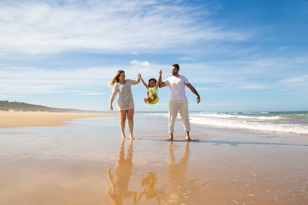Веселые родители и маленькая девочка наслаждаются прогулками и занятиями на пляже, ребенок, держащий руки родителей, прыгает и вскидывает ноги