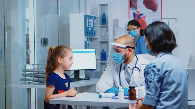 Cheerful pediatrician smiling at little girl during medical visit. Specialist in medicine with protection mask providing health care services, consultation, treatment, examination in hospital cabinet.