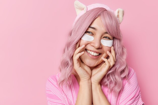 Cheerful pink haired Asian woman keeps hands on face smiles gladfully applies white beauty patches under eyes wears headband dressed in casual clothes isolated over pink background blank space
