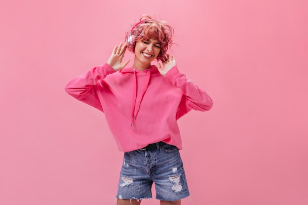 Веселая розоволосая женщина в джинсовых шортах и огромном худи слушает музыку в наушниках и танцует на изолированной стене