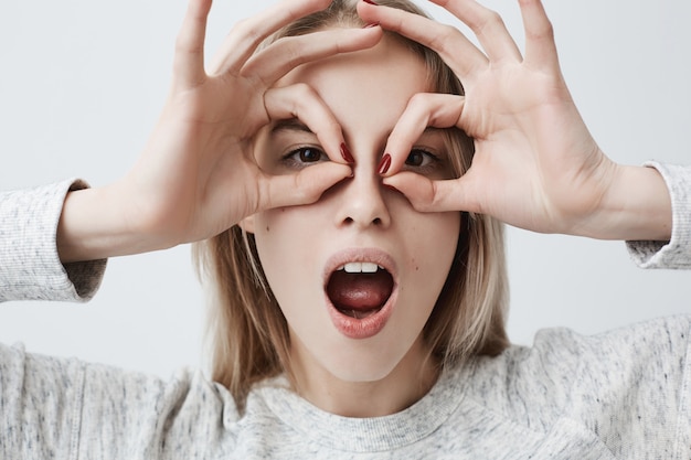 Foto gratuita giovane femmina allegra allegra con capelli biondi che mostra i gesti giusti con entrambe le mani, fingendo di indossare gli occhiali, sorridendo ampiamente, godendo della sua spensierata vita felice. emozioni e sentimenti positivi.