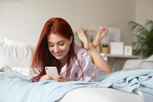 Веселая игривая молодая рыжая женщина в пижаме, лежащая на кровати в своей комнате с поднятыми ногами, радостно улыбаясь, флиртуя со своим парнем, отправляя ему текстовое сообщение онлайн с помощью мобильного телефона