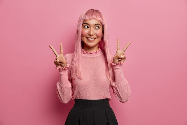 Cheerful pleasant looking woman with long pink hair, shows victroy or peace gesture, smiles joyfully and feels happy, looks aside, being in good mood