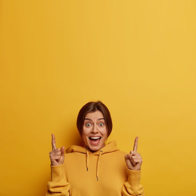 Free photo cheerful pleased caucasian woman points above, has glad expression, shows nice offer, gives recommendation going upstairs, wears comfortable yellow sweatshirt, involved in advertising campaign