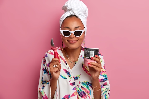  cheerful pleased dark skinned woman enjoys pleasant taste of strawberry ice cream, holds spoon for eating, spends summer vacation at home, isolated on pink wall. Frozen dessert