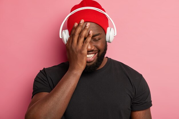 Cheerful pleased guy in red hat, closes eyes, smiles broadly, chooses song for relaxation wears stereo headphones, isolated on pink wall, spends free time listening music.