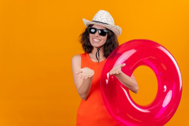 A cheerful and pleased young woman with short hair in an orange shirt wearing sun hat and sunglasses holding inflatable ring 