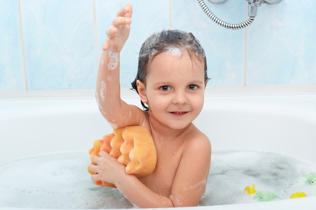 Cheerful positive adorable small child taking bath and washing herself with yellow sponge