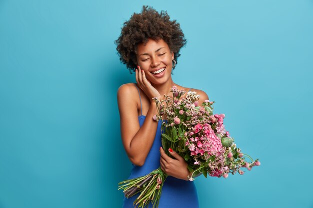 Cheerful positive dark skinned young woman smiles happily with closed eyes holds big bouquet of flowers