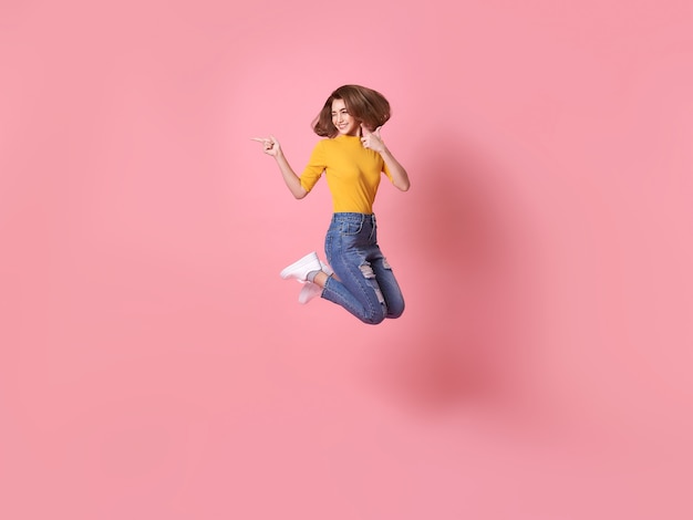 Cheerful positive girl jumping in the air with raised hand pointing to copy space isolated on pink background.