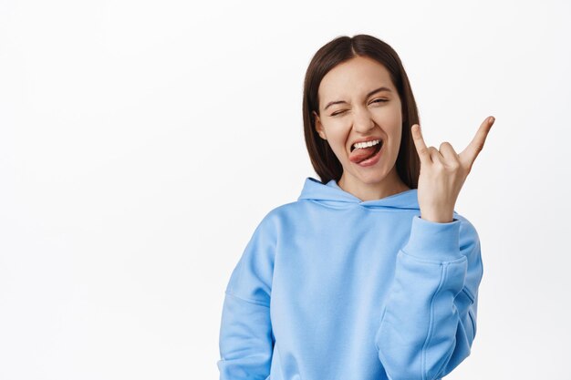Cheerful and positive girl shows horns heavy metal sign, winking and showing tongue, having fun, enjoy event and celebrating, standing in hoodie against white background.